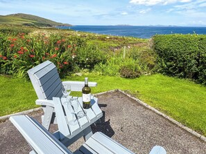 Un banc en bois, une bouteille de vin et des verres, disposés sur un chemin de gravier offrant une vue sur la mer et les collines.