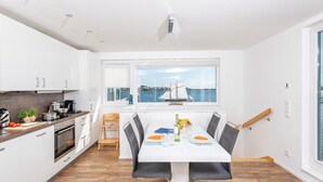 A modern kitchen with white cabinets, a dining area with a white table and chairs, and a view of the sea through a window. - Lovely ship in Olpenitz with sauna (Olpenitz)