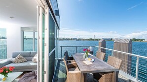 A balcony with a sofa, a table with chairs, and a view of the water and distant buildings. - Lovely ship in Olpenitz with sauna (Olpenitz)