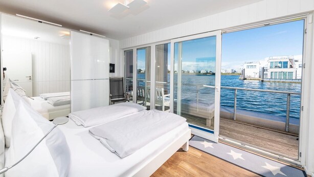 A modern bedroom with a large bed, a sliding glass door leading to a balcony with a view of the water and buildings. - Lovely ship in Olpenitz with sauna (Olpenitz)