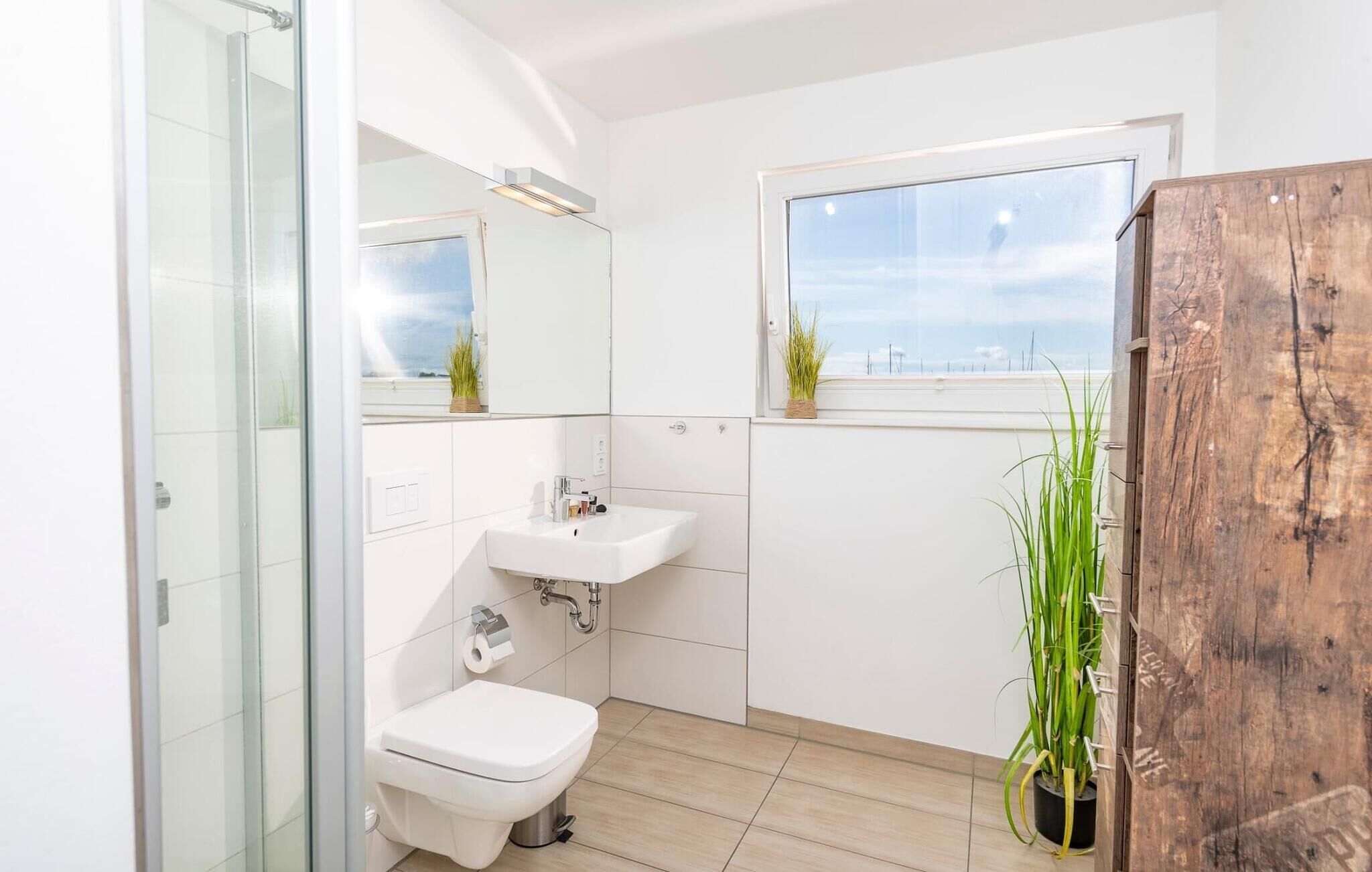 Um banheiro moderno com box de vidro, pia branca e vaso sanitário. Há uma janela com vista para o céu e uma planta no parapeito.