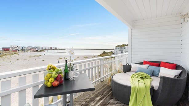 Un balcon avec un canapé en osier, une table avec des fruits et une bouteille, et une vue sur la plage et les bâtiments.
