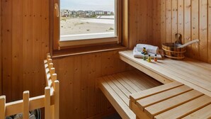 Un sauna avec des bancs en bois, une fenêtre donnant sur une plage et des bâtiments, ainsi qu’une serviette et des bouteilles posées sur une étagère.