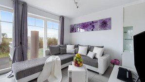Un salon moderne avec un canapé gris, des coussins blancs, une petite table blanche sur laquelle sont disposés des fruits, et une grande fenêtre avec des rideaux.