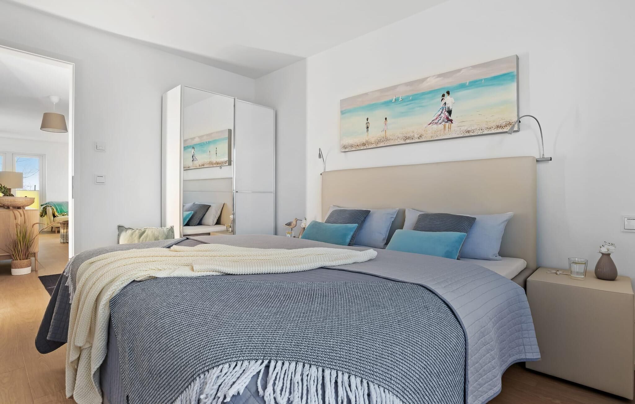 Une chambre à coucher avec un lit, des oreillers, une table de chevet, une lampe et un tableau représentant une scène de plage.