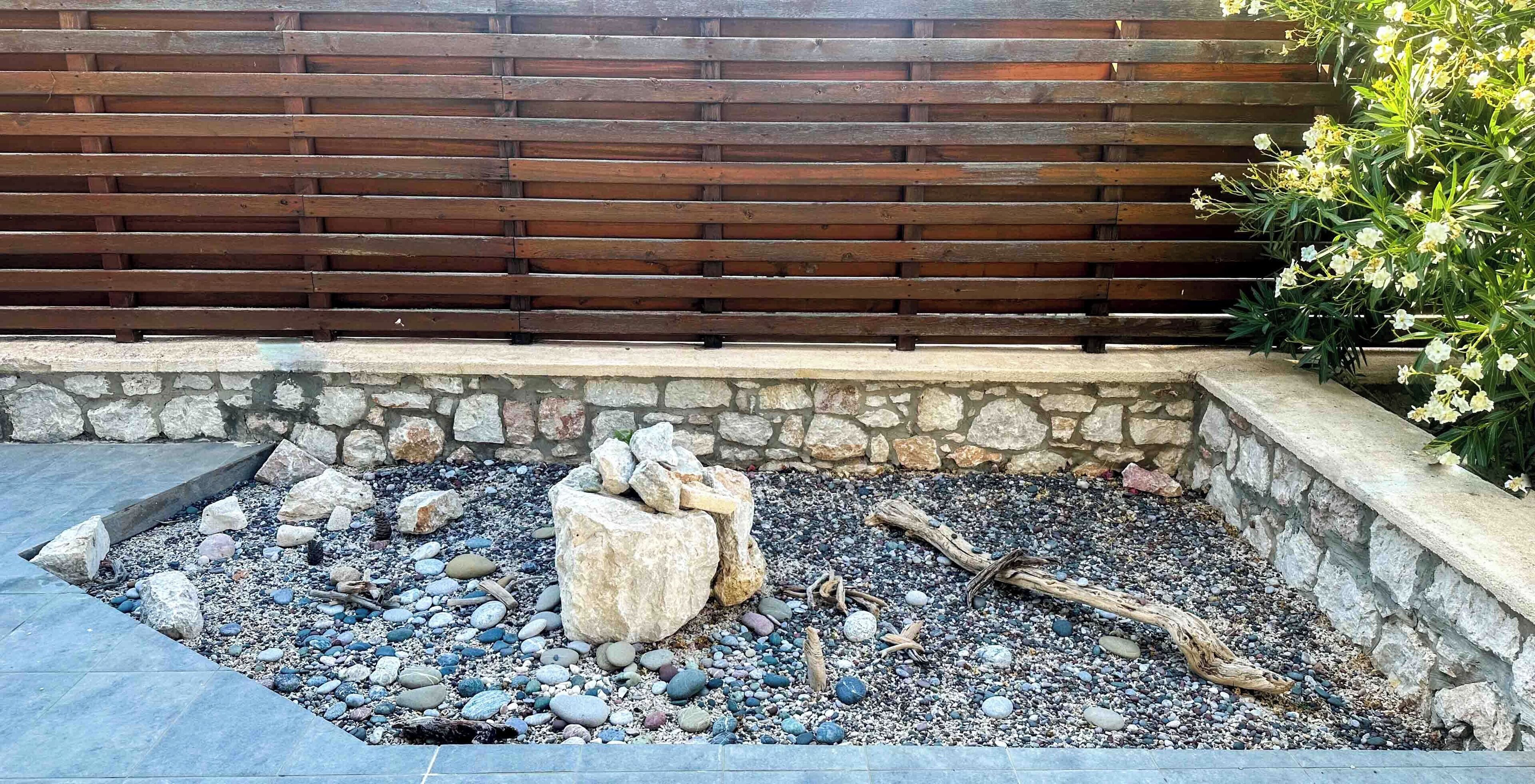 A stone wall with a wooden fence, a gravel area with rocks and logs, and a plant with white flowers.