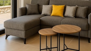 Un salon moderne avec un canapé d’angle gris, deux petites tables rondes en bois et un coussin jaune.