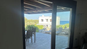 Une pièce dotée d’une porte-fenêtre coulissante donnant sur une terrasse extérieure avec une table et des chaises, une vue sur la mer et un bâtiment en arrière-plan.