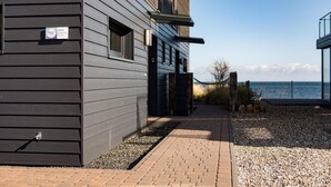 A modern building with a grey exterior, a pathway leading to a gated entrance, and a view of the sea. - Cozy home in Olpenitz with sauna (Olpenitz)
