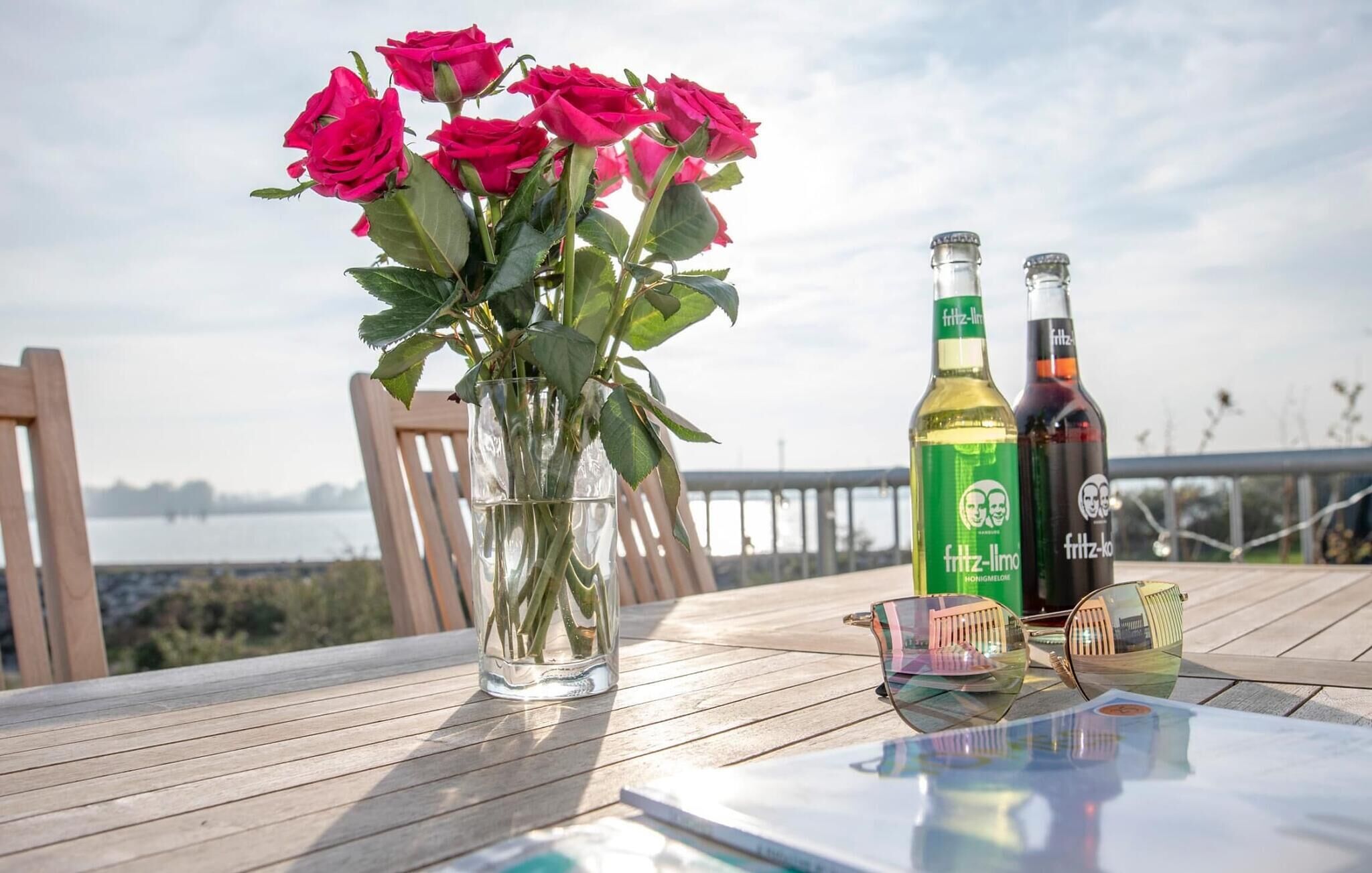 A table with a vase of pink roses, two bottles of Fritz-Limo alcohol, and a pair of sunglasses on a wooden deck with a view of a body of water.