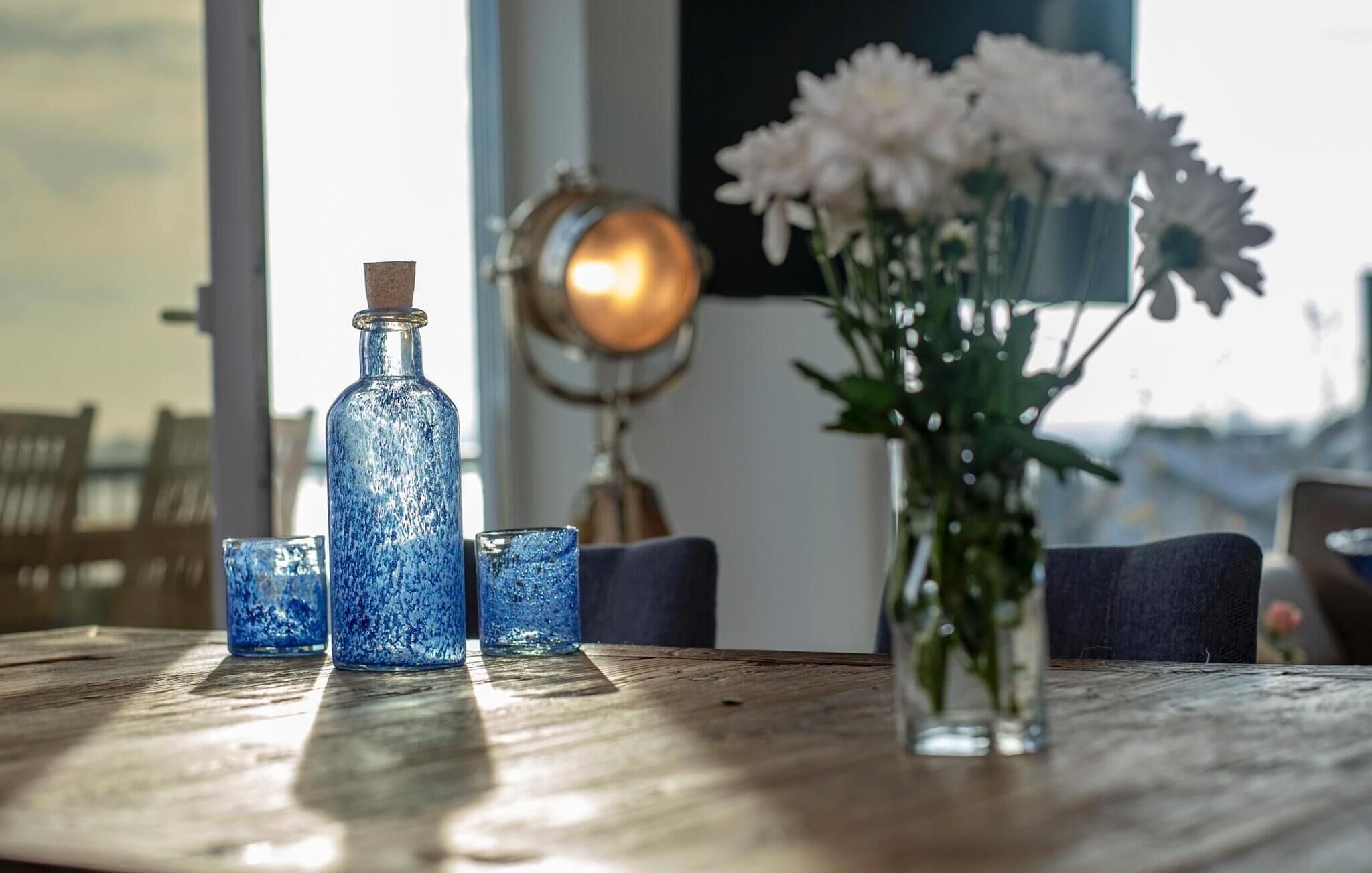 Una mesa de madera con una botella de vidrio azul, dos vasos pequeños y un florero con flores blancas.