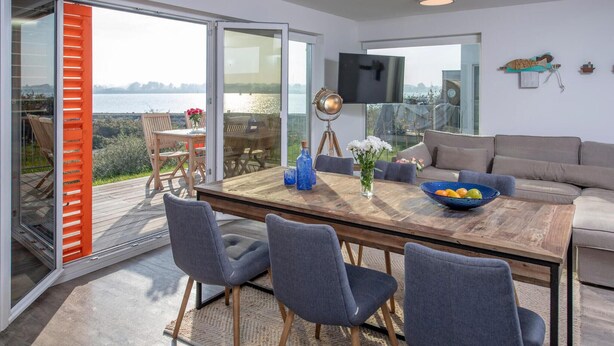 Una sala de estar moderna con una mesa de comedor de madera, sillas tapizadas en gris, un sofá gris y un ventanal con vistas a un cuerpo de agua.
