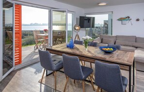 A modern living room with a wooden dining table, grey upholstered chairs, a grey sofa, and a large window overlooking a body of water. - Nice apartment in Olpenitz with sauna (Olpenitz)