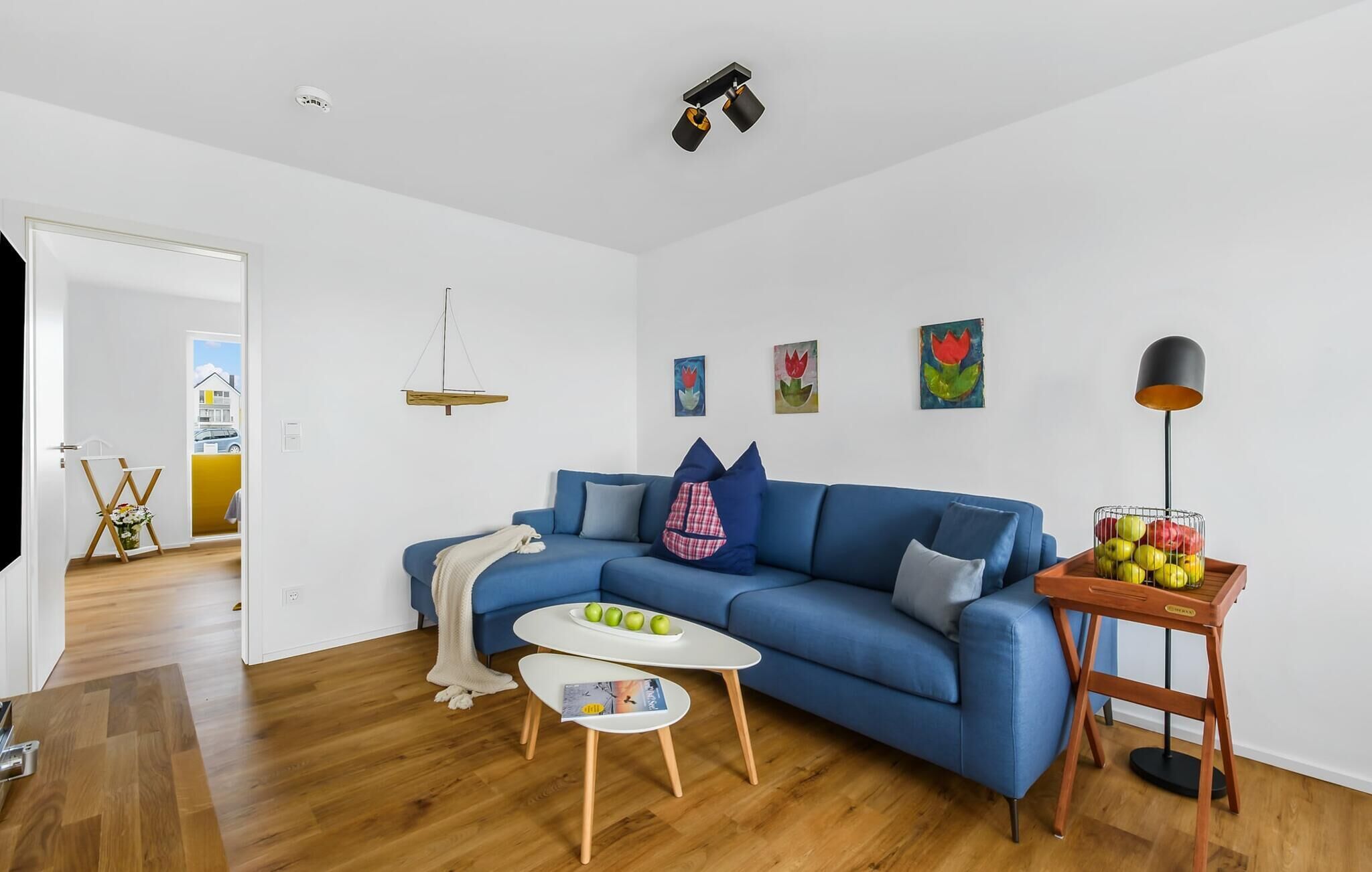 Een moderne woonkamer met een blauwe bank, een klein wit tafeltje en een houten bijzettafeltje met fruit.
