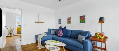 Een moderne woonkamer met een blauwe bank, een klein wit tafeltje en een houten bijzettafeltje met fruit.