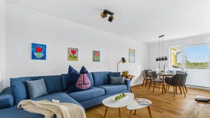 A modern living room with a blue sofa, a small white table, and a dining area with wooden chairs and a table. There are wall-mounted artworks and a window with a view. - 2 bedroom beach front home in Olpenitz (Olpenitz)