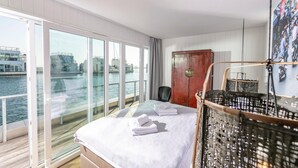 Ein Hotelzimmer mit großem Fenster und Blick auf Wasser und Gebäude, ein Bett mit weißer Bettwäsche, eine Holztür und ein Korbwäscheskorb.