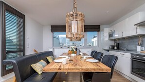 Eine moderne Küche mit weißen Schränken, einem Holzespeisetisch, der für sechs Personen gedeckt ist, und einem schwarzen Ledersofa mit gemusterten Kissen.