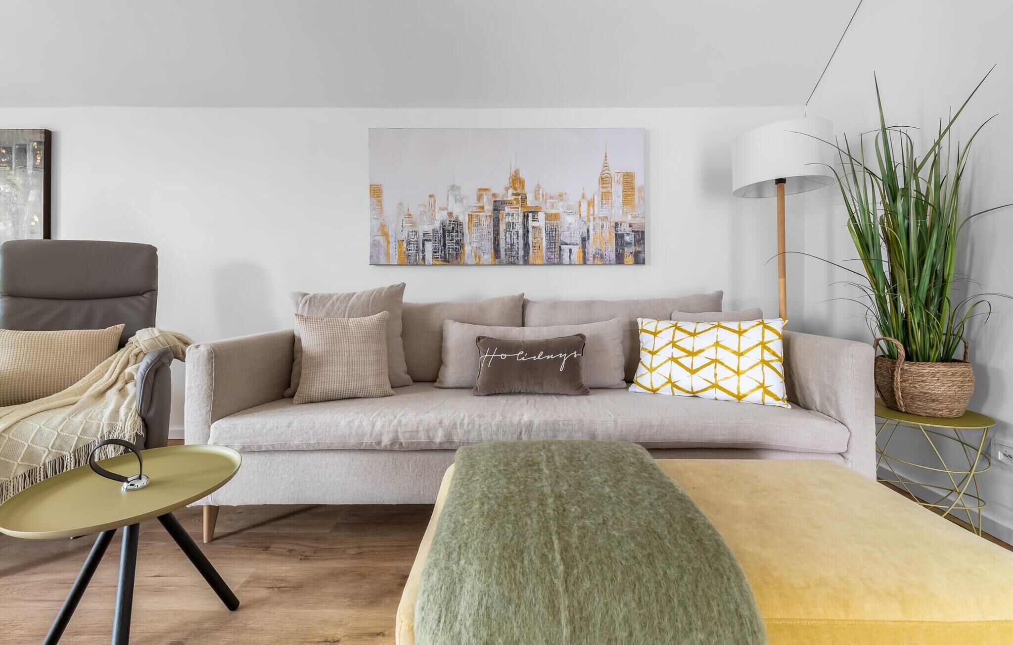 A living room with a sofa, a small round table, a green ottoman, and a cityscape painting on the wall.