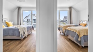 Een moderne slaapkamer met twee bedden, houten vloeren en grote ramen met uitzicht op omliggende huizen.