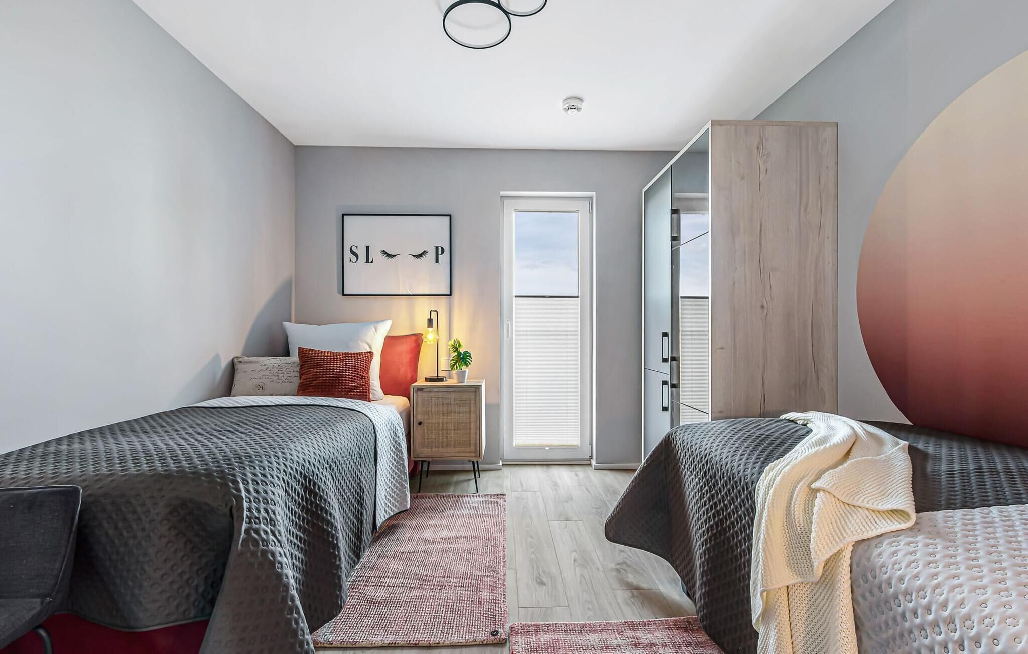 Une chambre moderne avec deux lits, une table de chevet avec une lampe, une œuvre d’art encadrée et une porte coulissante donnant sur un placard.