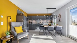Una cocina moderna con gabinetes grises, un comedor con mesa de madera y sillas grises, y un sofá con un cojín amarillo.