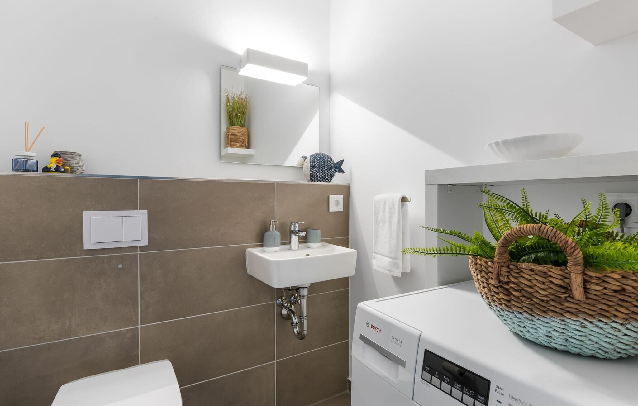 Casa de banho moderna com lavatório branco, espelho e máquina de lavar Bosch. Há uma toalha pendurada na parede e uma cesta com plantas no balcão.