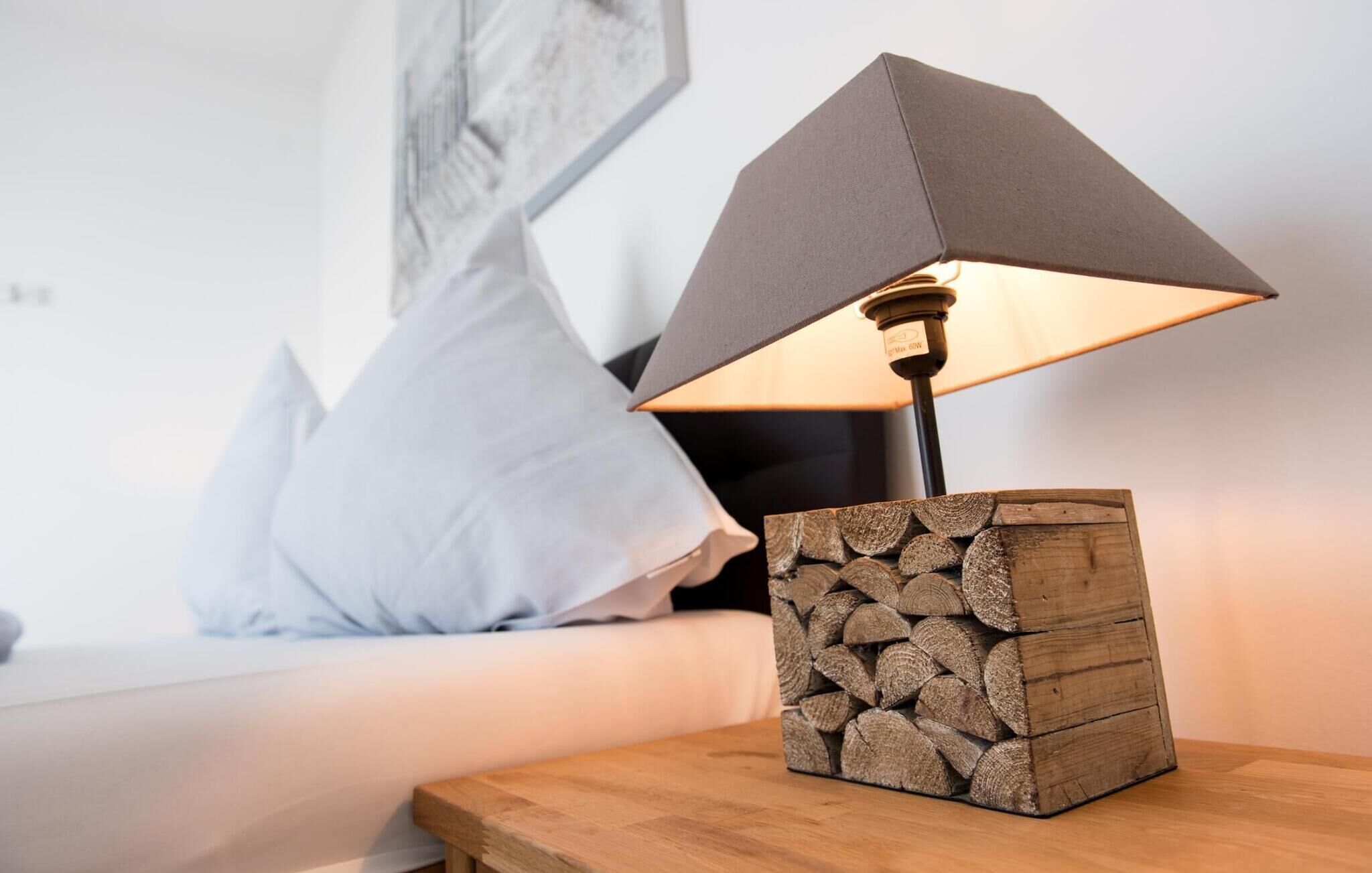 A bedside table with a lamp, a stack of wood logs, and a white sofa with a pillow.