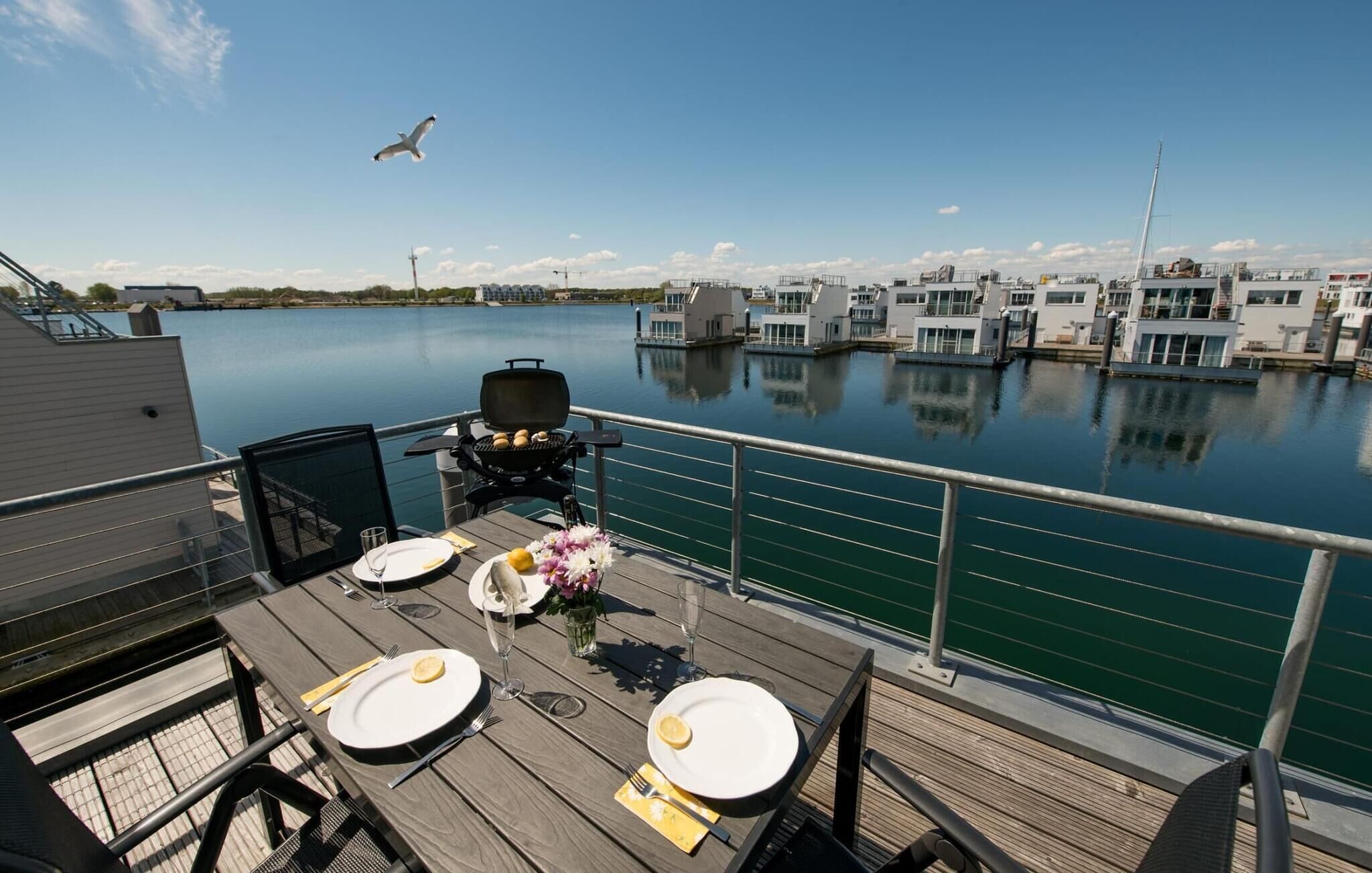 Een eethoek met uitzicht op een jachthaven, een grill en een gedekte tafel voor twee met borden en bestek.