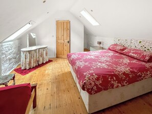Une chambre avec un plancher en bois, un lit recouvert d’une courtepointe à motifs floraux, une petite table avec une nappe à motifs floraux, une porte en bois et un puits de lumière.
