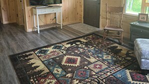 A room with wooden walls, a television on a white stand, a patterned rug, a wooden chair, and a window with a view of trees. - Right on the Tuscobia Trail Cozy 2-bedroom cabin in  Winter with WiFi,  AC  (Winter)