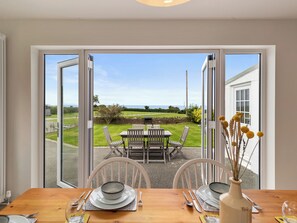 Un coin repas avec une table dressée pour deux personnes, une vue sur le jardin à travers des portes vitrées et un bâtiment blanc en arrière-plan.