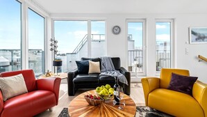 Ein modernes Wohnzimmer mit einer schwarzen Couch, einem roten Sessel und einem gelben Sessel. Ein Holztisch bietet Platz für Früchte und ein Weinglas. Große Fenster bieten Blick auf die Stadt.