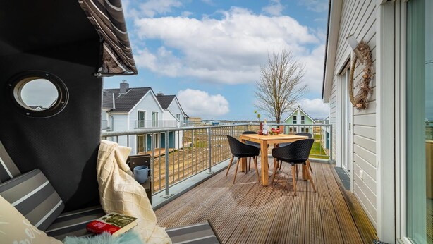A balcony with a wooden table and chairs, a view of houses, and a clear sky.