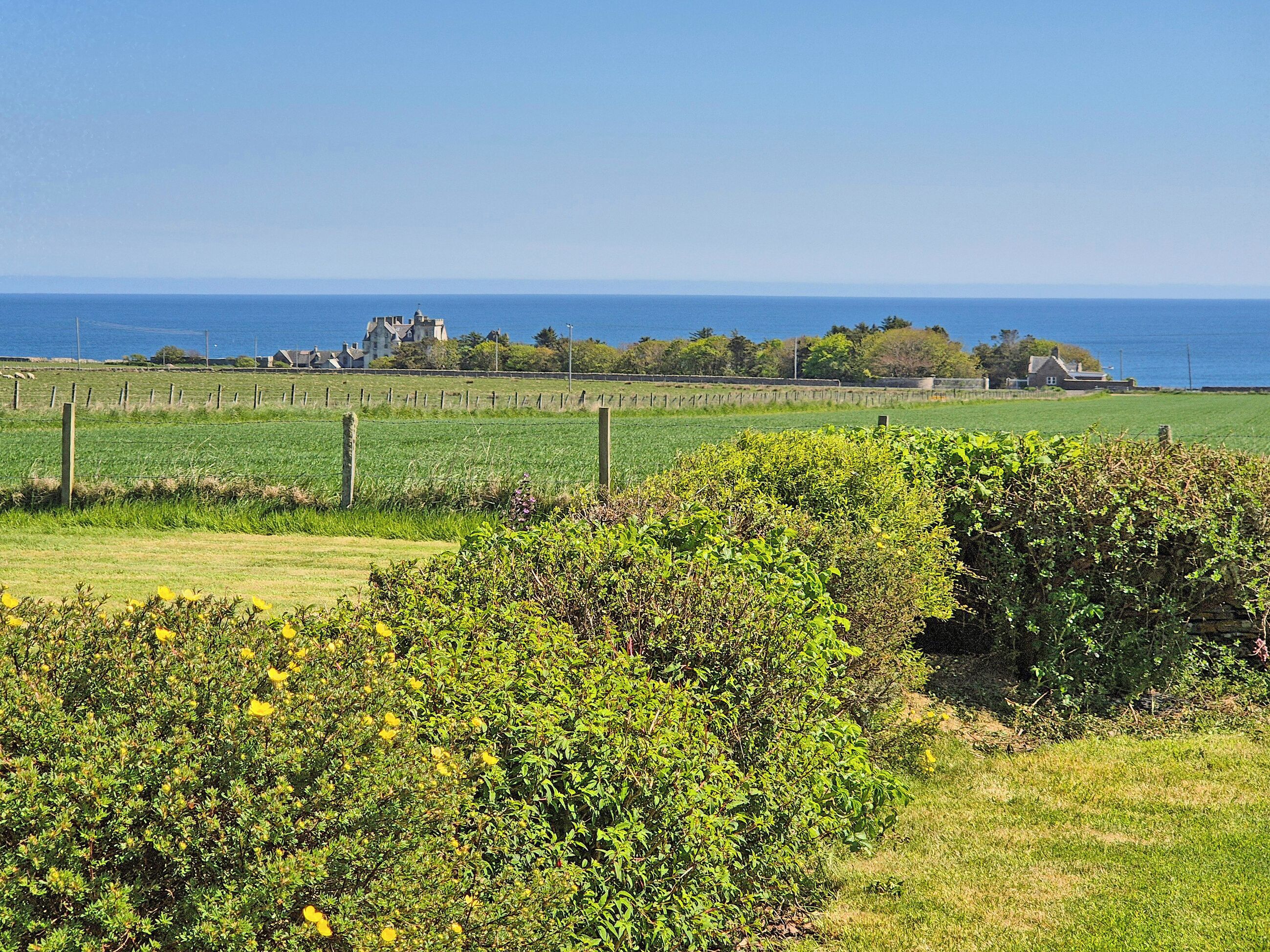 Un paisaje costero con un campo, una siega y una casa distante al mar.