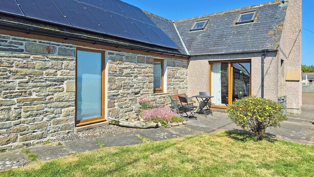 A stone house with a solar panel roof, a stone chimney, and a small garden with a bench and table. - 2 bedroom accommodation in North Keiss, Caithness (North Keiss, Caithness)