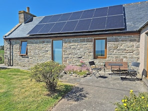 A stone building with a solar panel roof, a window, and a patio with chairs and a table.