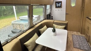 Interior acogedor de una caravana con mesa de comedor, asientos acolchados, una planta pequeña y ventana con vistas a una zona verde.