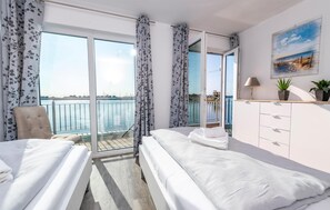Habitació d'hotel amb dos llits, un balcó que dona al mar, un quadre a la paret i una petita planta al taulell.