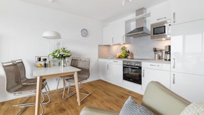 A modern kitchen with white cabinetry, a built-in oven, a microwave, and a sink. There is a dining area with a table and chairs, and a clock on the wall.