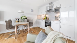 A modern kitchen with white cabinets, a built-in oven, and a microwave. There is a dining area with a table and chairs, and a clock on the wall.