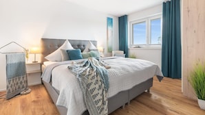 A modern bedroom with a large bed, a bedside table with a lamp, a window with curtains, and a wooden floor.