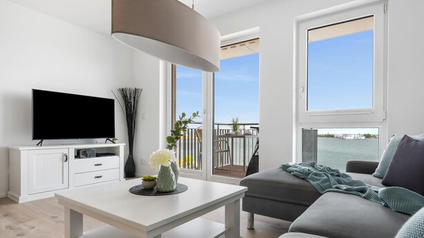 A modern living room with a flat-screen TV, white furniture, a balcony with a view of the water, and a large window. - Nice apartment in Olpenitz with WiFi (Olpenitz)