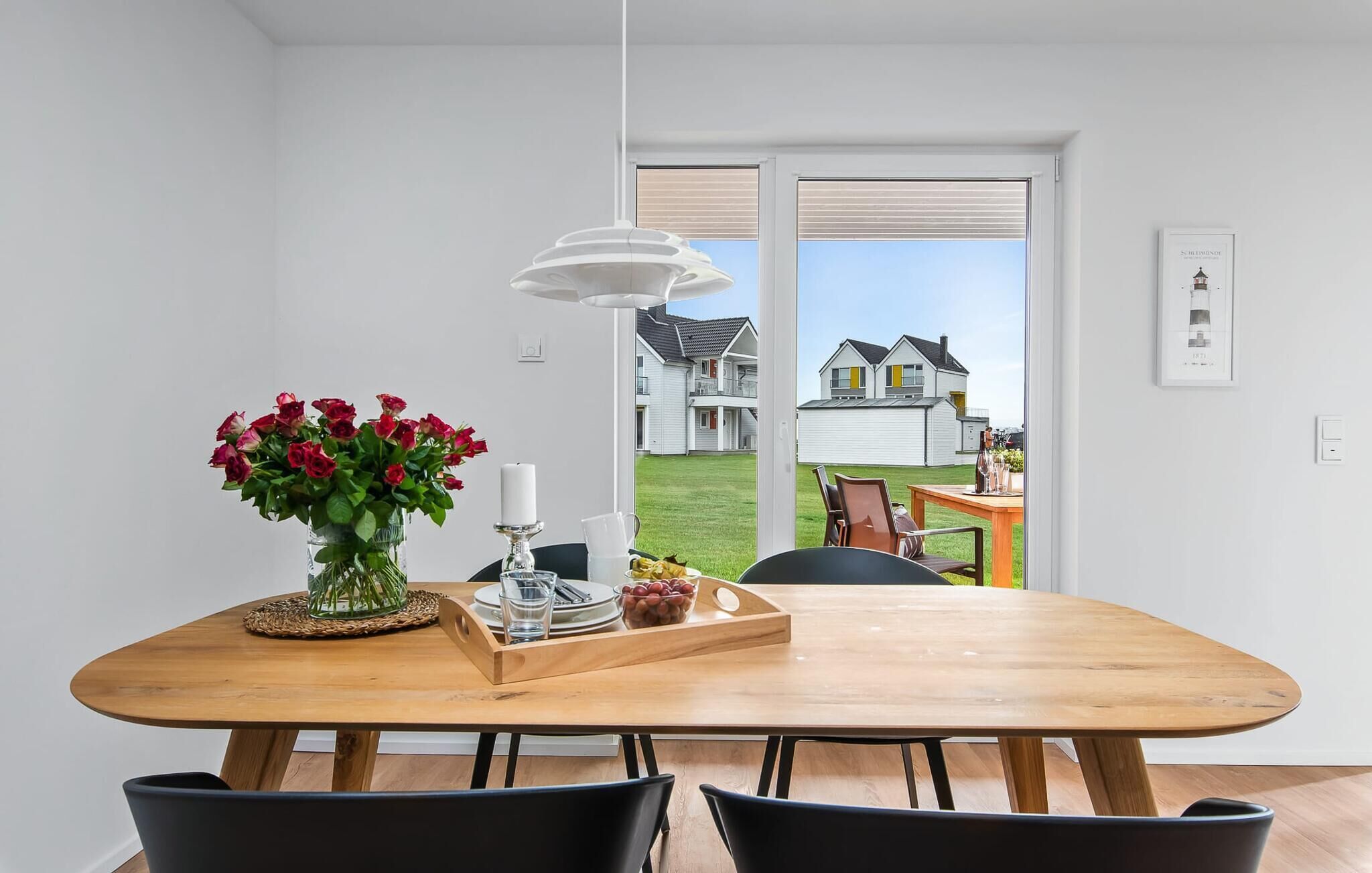 Een eethoek met een houten tafel, zwarte stoelen, een vaas rode bloemen, een kaars en een dienblad met fruit en glazen.
