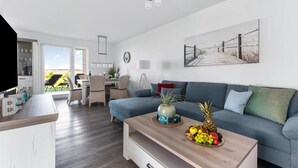 A modern living room with a blue sofa, a wooden coffee table with fruit, and a dining area with wicker chairs. - Beach front apartment in Olpenitz (Olpenitz)