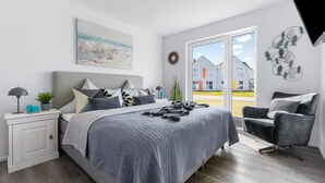 A modern bedroom with a large bed, a bedside table with a lamp, a potted plant, and a view of a residential area through a sliding door. - Beach front apartment in Olpenitz (Olpenitz)