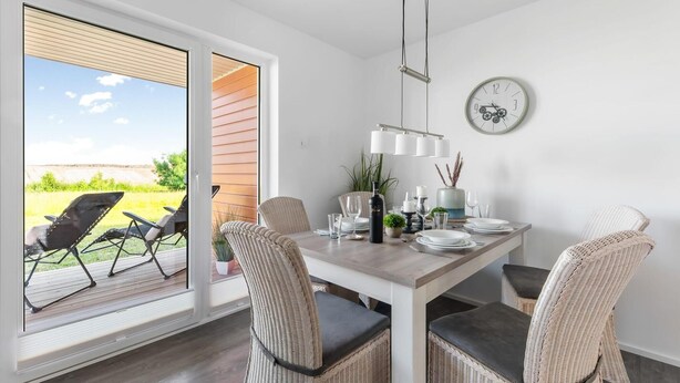 A dining area with a table set for four, wicker chairs, a clock on the wall, and a view of the outdoors through a sliding door. - Beach front apartment in Olpenitz (Olpenitz)