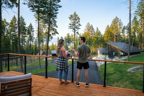 Um deck de madeira com guarda-corpo, vista para uma área arborizada e um pequeno edifício.