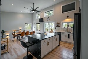 A modern kitchen with a large island, white cabinetry, and a black countertop. There are multiple windows providing natural light and views of the outdoors. - Flathead Lake View: Luxe Estate w/ Pool & Hot Tub (Lakeside)