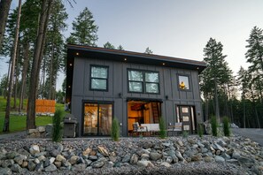 A modern two-story house with a stone foundation, large windows, and a patio area with outdoor furniture.